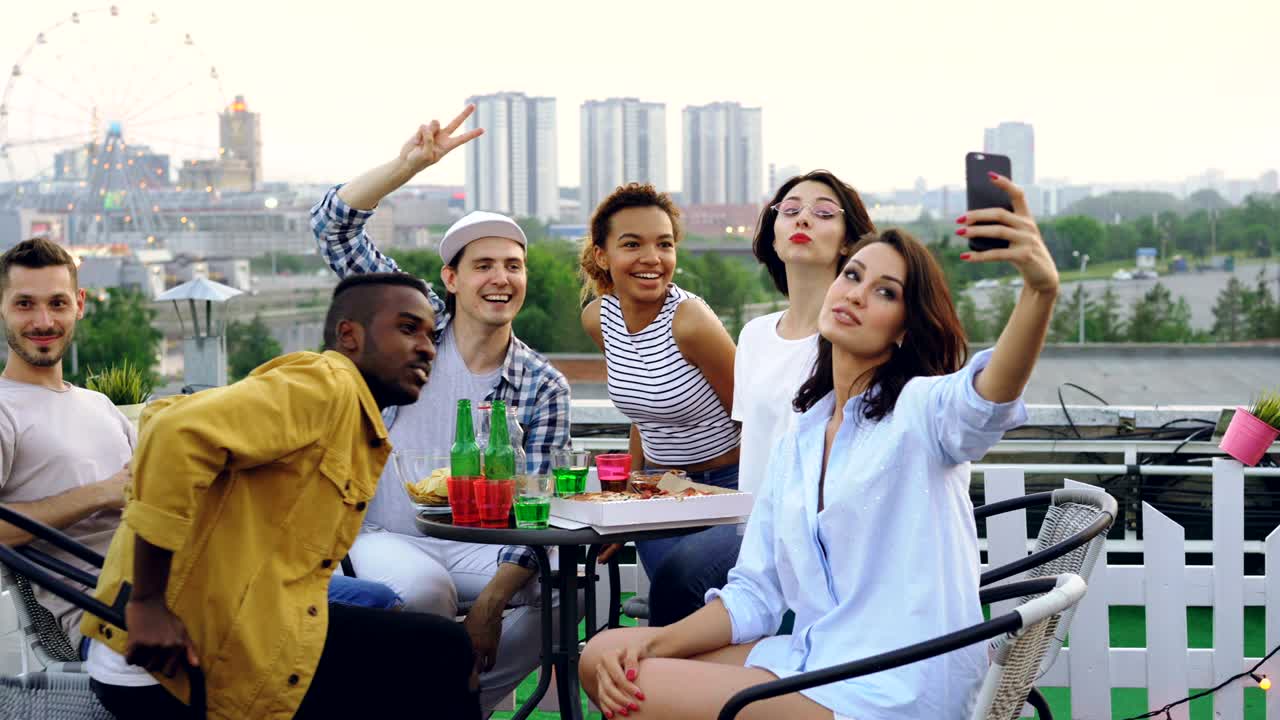 una joven bonita está sosteniendo un teléfono inteligente y tomando selfies con amigos sentados en la mesa con comida y bebidas en el techo. la hermosa ciudad está en el fondo.