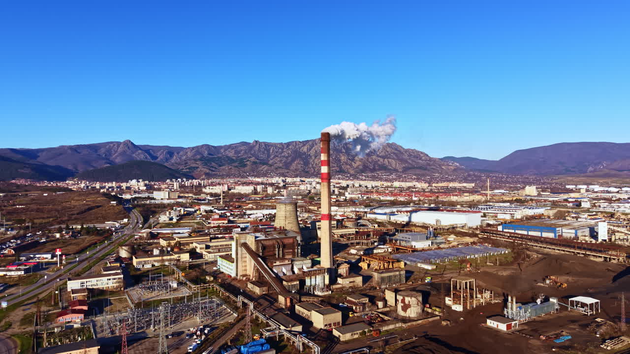 Área industrial com fábrica e chaminé sob céu azul claro em região montanhosa.