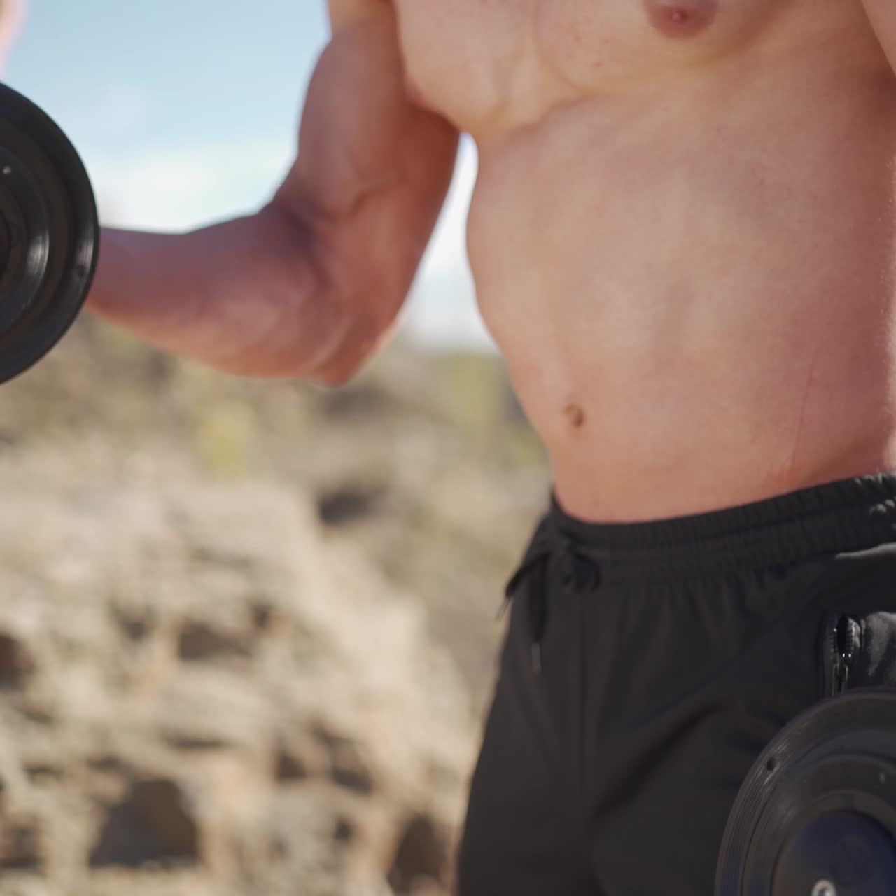 Healthy bodybuilder with heavy dumbbells. Muscular sportsman lifting metal dumbbells outdoors. Physical workout in nature. Close-up.