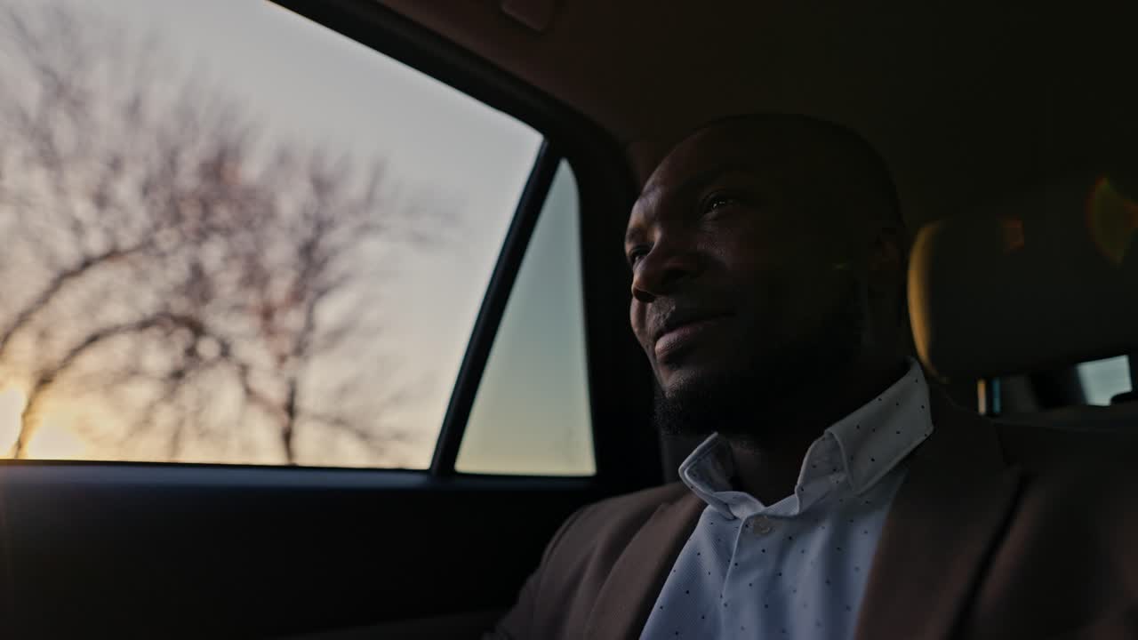 갈색 슈트를 입은 행복한 검은 피부 사업가는 일몰 동안 일하는 힘든 하루를 마치고 저녁에 차로 집으로 돌아옵니다. 검은 피부를 가진 남자는 차를 운전하고 태양과 일몰의 반이는 광선을 창문 밖으로 바라보고 있습니다.