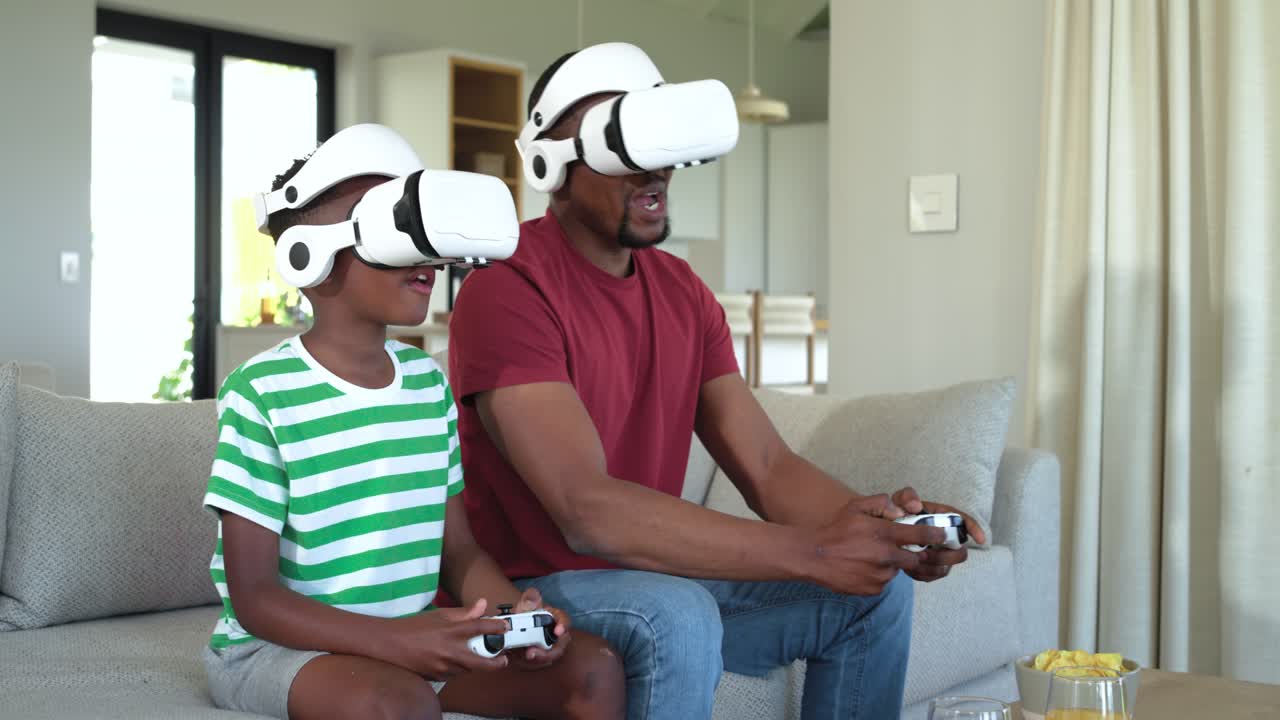 Padre afroamericano e figlio con visori VR e controller che reagiscono all'inizio di un gioco sul divano