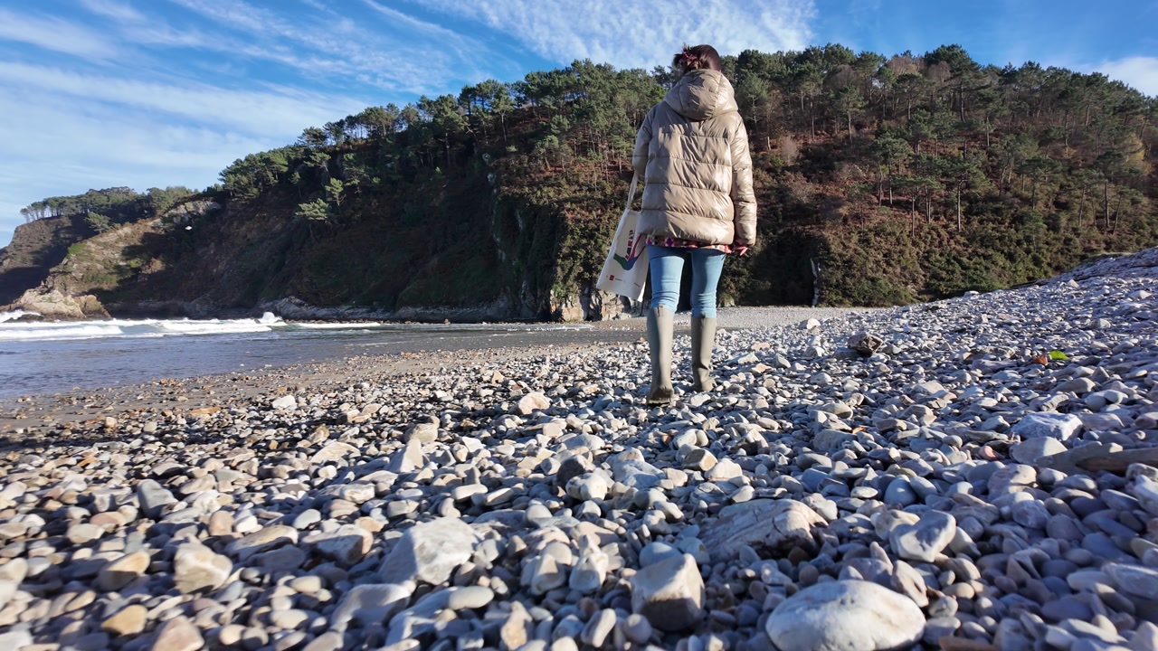Woman standing on pebble beach facing ocean waves and cliffs