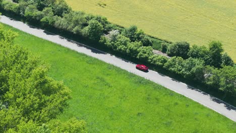 tracking drone shot of red car moving on countryside road on sunny summer day, between meadows, trees and railway