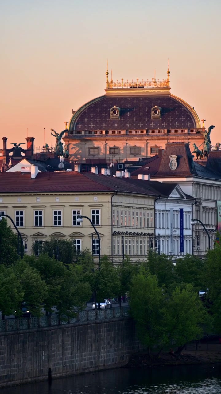 Vista verticale della cupola e della cresta dorata del tetto del Teatro Nazionale sopra gli edifici lungo il fiume al crepuscolo, Praga, Repubblica Ceca