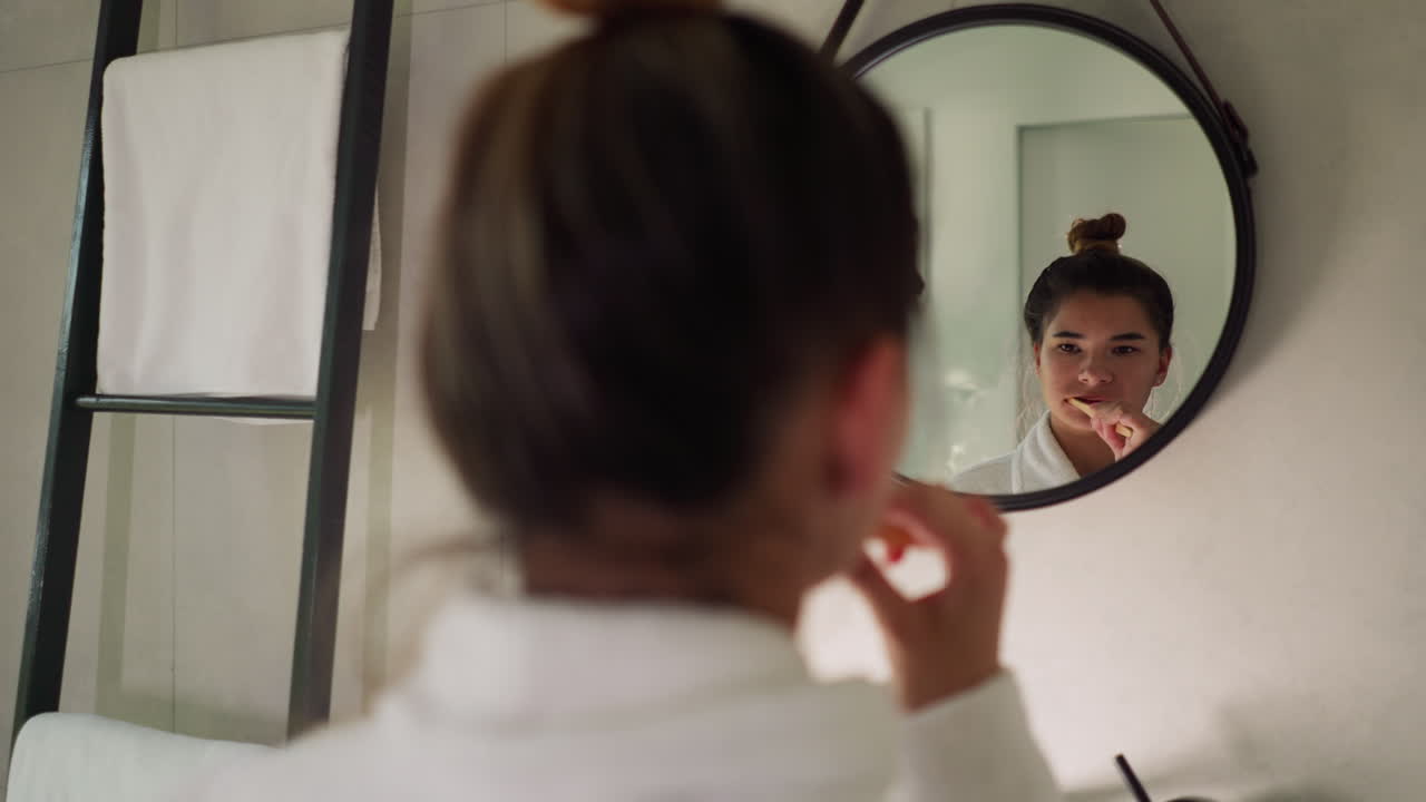 Woman brushes teeth by mirror at home. Pretty young lady in bathrobe does oral cavity hygienic procedure looking in bathroom. Everyday routine