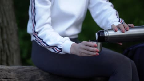 vista panorámica de una joven caucásica sentada en un enorme bosque de troncos y sirviéndose una bebida caliente de una botella de termo