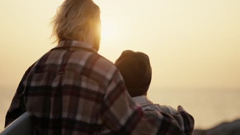 vista trasera de un feliz chico rubio en una camisa a cuadros y su novia rubia en un sombrero negro y camisa a cuadrados de pie y abrazando y mirando al mar por la mañana al amanecer
