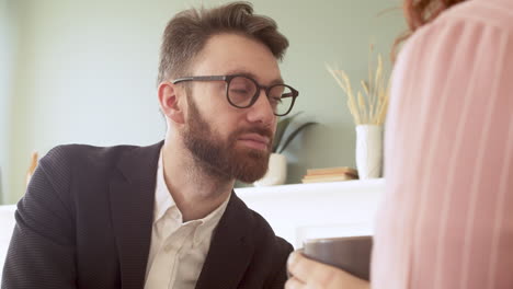 el primer plano de un joven con barba y anteojos escuchando a alguien hablando mientras está sentado en una sala de estar moderna