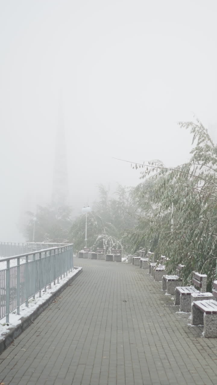 Winter cityscape with thick fog and fresh snowfall blanketing park benches, metal railings, and quiet walkway framed by light poles and frosted trees, creating calm muted serene urban scenery