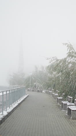winter cityscape with thick fog and fresh snowfall blanketing park benches, metal railings, and quiet walkway framed by light poles and frosted trees, creating calm muted serene urban scenery