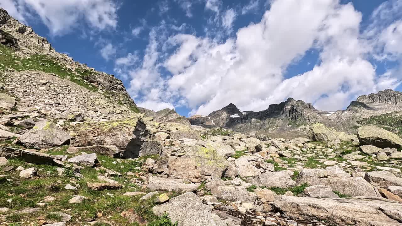 A rugged high-altitude landscape of loose boulders and alpine grass in the Lepontine Alps, Piedmont, Italy, set beneath craggy peaks and a sky filled with drifting summer clouds