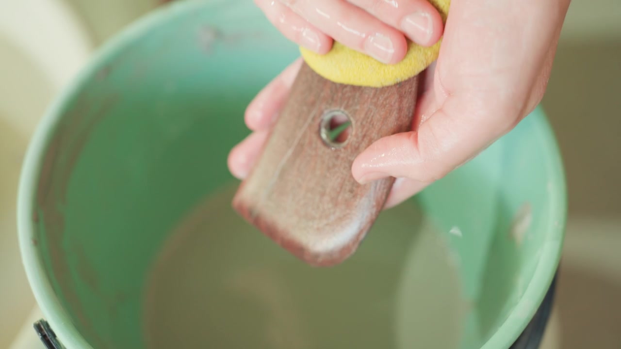 Artisan rinçant une nervure en bois dans une bassine verte, gros plan des mains et de l'éponge. Scène de studio d'inspection finale après nettoyage, gouttelettes d'eau, grain lisse, prêt à l'emploi, satisfaction tranquille