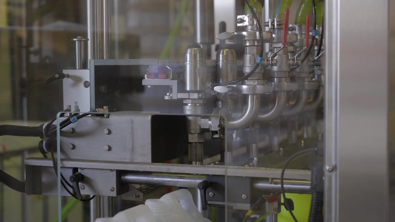 Industrial bottling machine with multiple nozzles behind a safety glass shield. Automated liquid filling process in a chemical production plant.