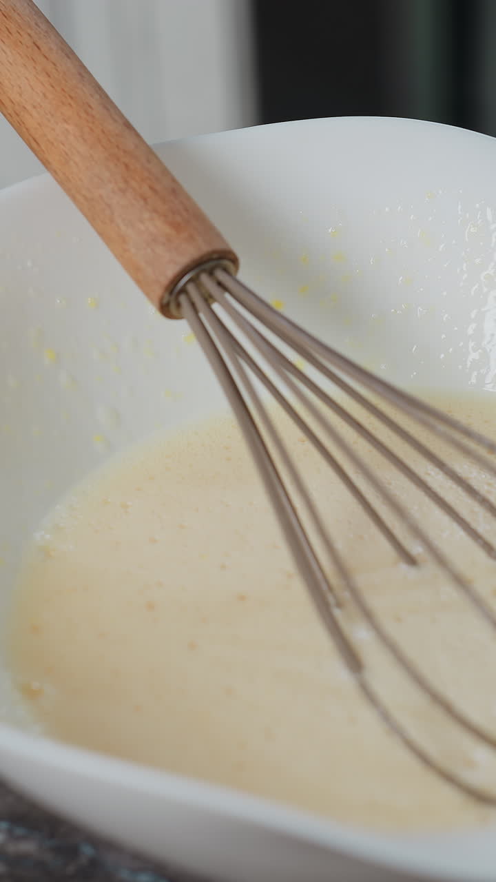 primer plano de un batidor descansando en la mezcla de horneado dentro de un cuenco blanco en la encimera, el fondo de la cocina presenta gabinetes modernos, diseño elegante y espacio de trabajo limpio