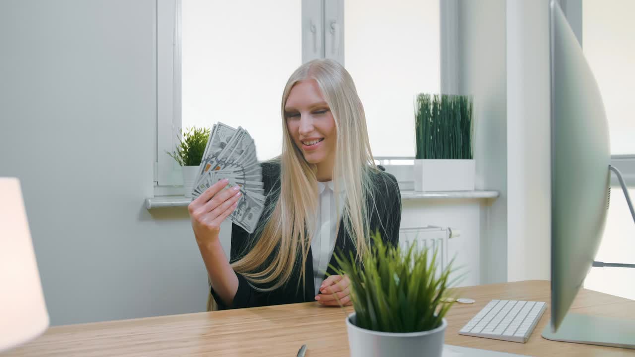 улыбающаяся бизнес-женщина с купюрой наличных. красивая счастливая блондинка сидит в офисе на рабочем месте с компьютером и держит вентилятор валютных банкнот и смотрит на камеру, улыбаясь.