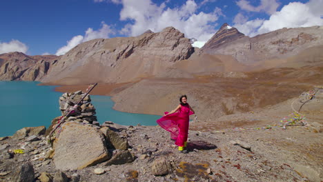 La-Actriz-Nepalí-Siente-El-Aire-En-El-Lago-Nepal-Tilicho,-El-Lago-De-Mayor-Altitud-Del-Mundo,-La-Toma-De-Un-Dron-Revela-El-Paisaje-De-La-Región-De-Annapurna,-Un-Vestido-Rosa-Haciendo-Alarde-De-La-Montaña-4k