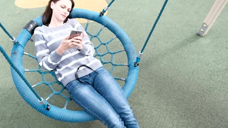 woman is swinging on empty webbed swing, typing and reading a message on mobile phone.