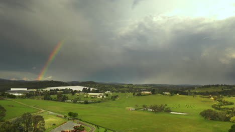 Aerial-view-moves-backward,-capturing-a-tranquil-natural-landscape-and-revealing-a-rainbow-among-heavy-clouds-in-the-sky