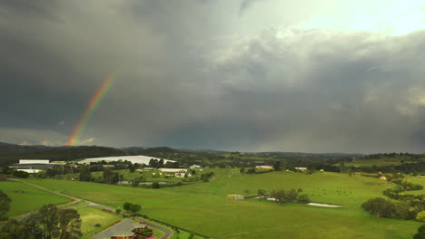 aerial view moves backward, capturing a tranquil natural landscape and revealing a rainbow among heavy clouds in the sky