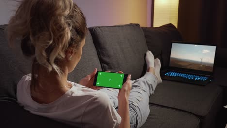 over the shoulder view woman lying on a sofa using phone with green mock-up screen chroma key watching content. horizontal phone green screen. using smartphone home.