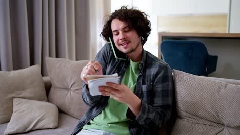 Confident-business-guy-brunette-with-curly-hair-communicates-on-the-phone-and-writes-down-important-things-in-a-conversation-on-a-notebook-at-home-on-the-sofa-in-a-modern-apartment