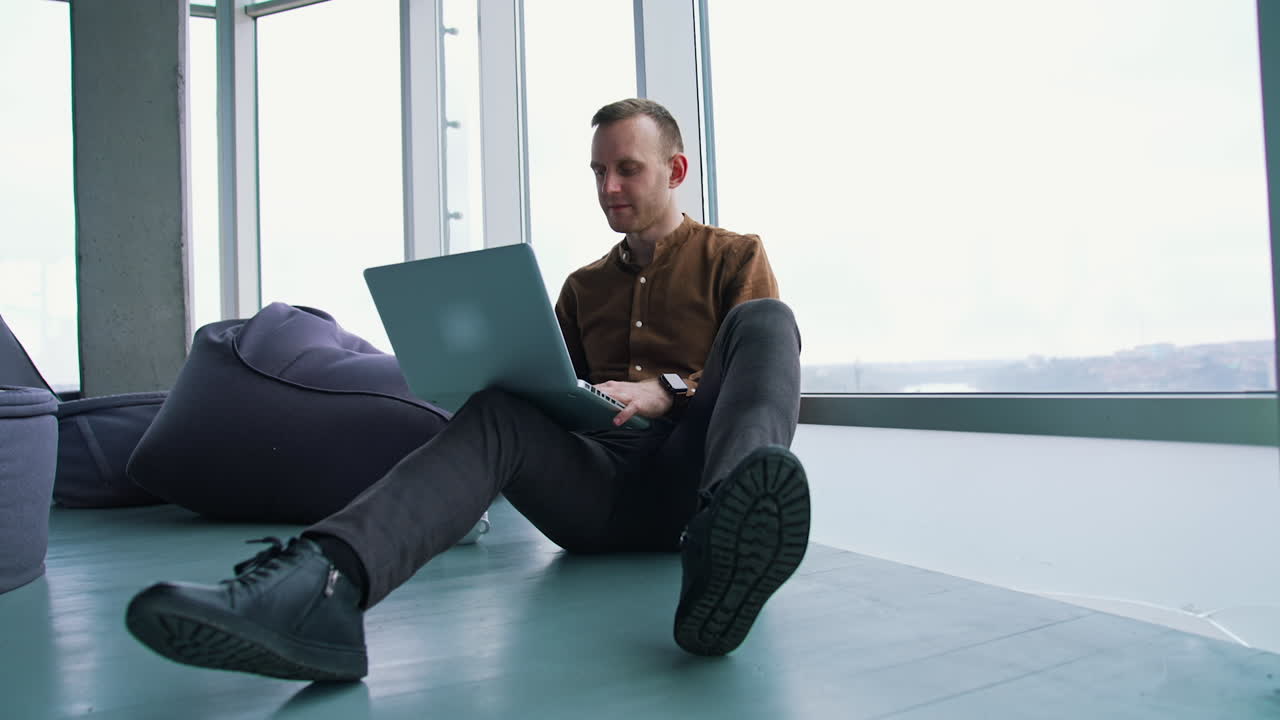 Young freelancer working on a laptop. Man in shirt and trousers sitting on a floor and using a gadget to work in office. Panoramic windows view.
