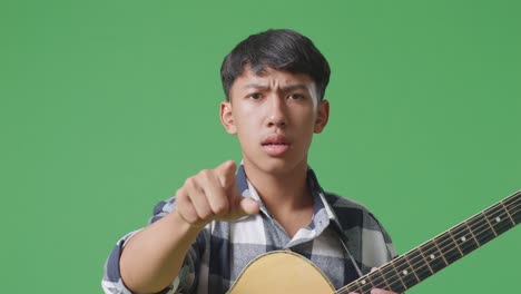 close up de un adolescente asiático con una guitarra gritando a la cámara mientras está de pie en el fondo de la pantalla verde