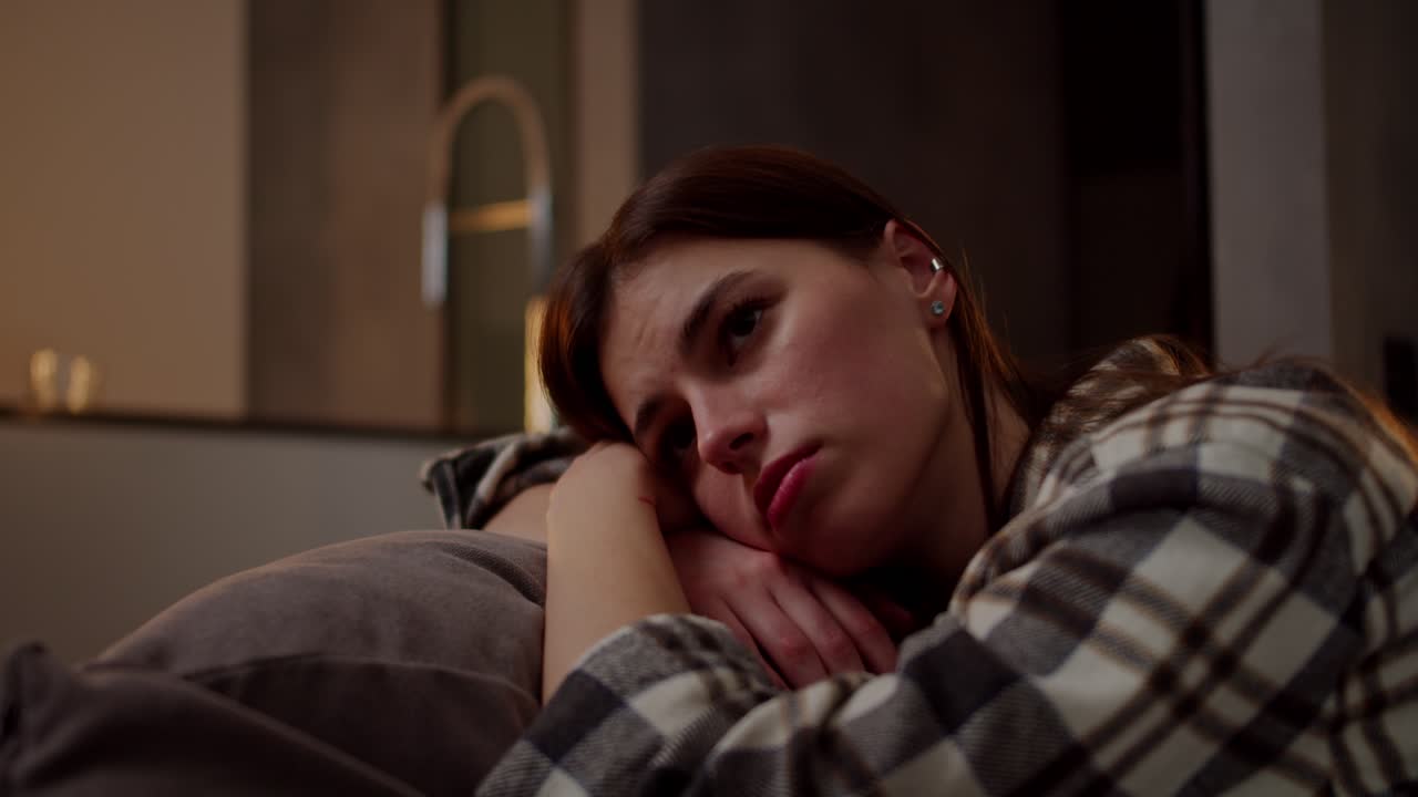 una tranquila y aburrida chica morena en una camisa a cuadros se apoya en una almohada en un sofá gris ella está aburrida y mira lejos en un apartamento moderno por la noche