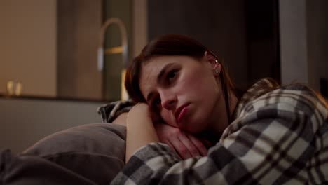 A-calm-and-bored-brunette-girl-in-a-checkered-shirt-leans-on-a-pillow-on-a-gray-sofa-she-is-bored-and-looks-away-in-a-modern-apartment-in-the-evening