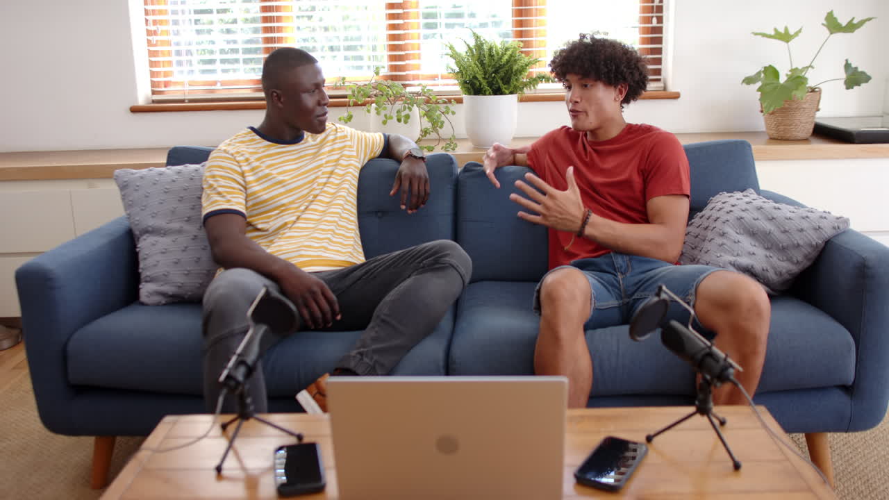 Recording podcast, two multiracial male friends on couch with microphones and laptop, at home
