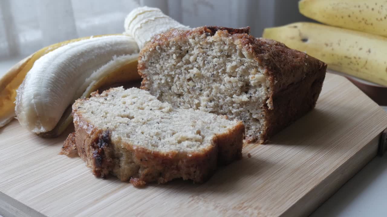 Freshly baked banana bread on a wooden cutting board with ripe bananas in the background