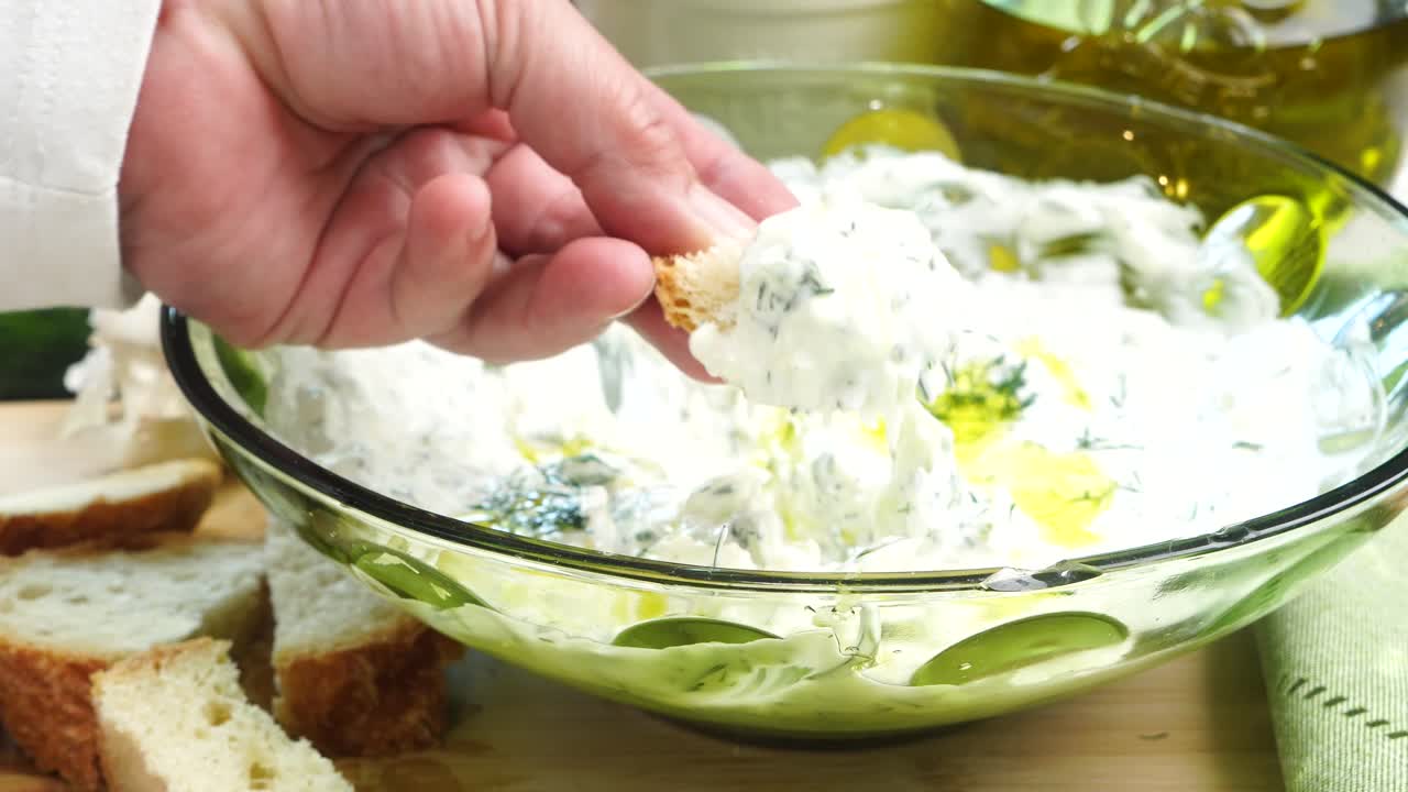 Dipping bread into Tzatziki sauce. Making Tzatziki Greek or Mediterranean sauce or dip for a party. Dip or sauce in a green bowl on a wooden cutting board with dill, green napkin and a grater.