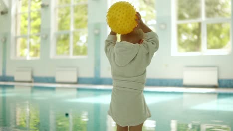 cute little boy throwing a yellow ball to the pool at the leisure center. swimming pool indoors. boy wears small white terry bathrobe. slow motion