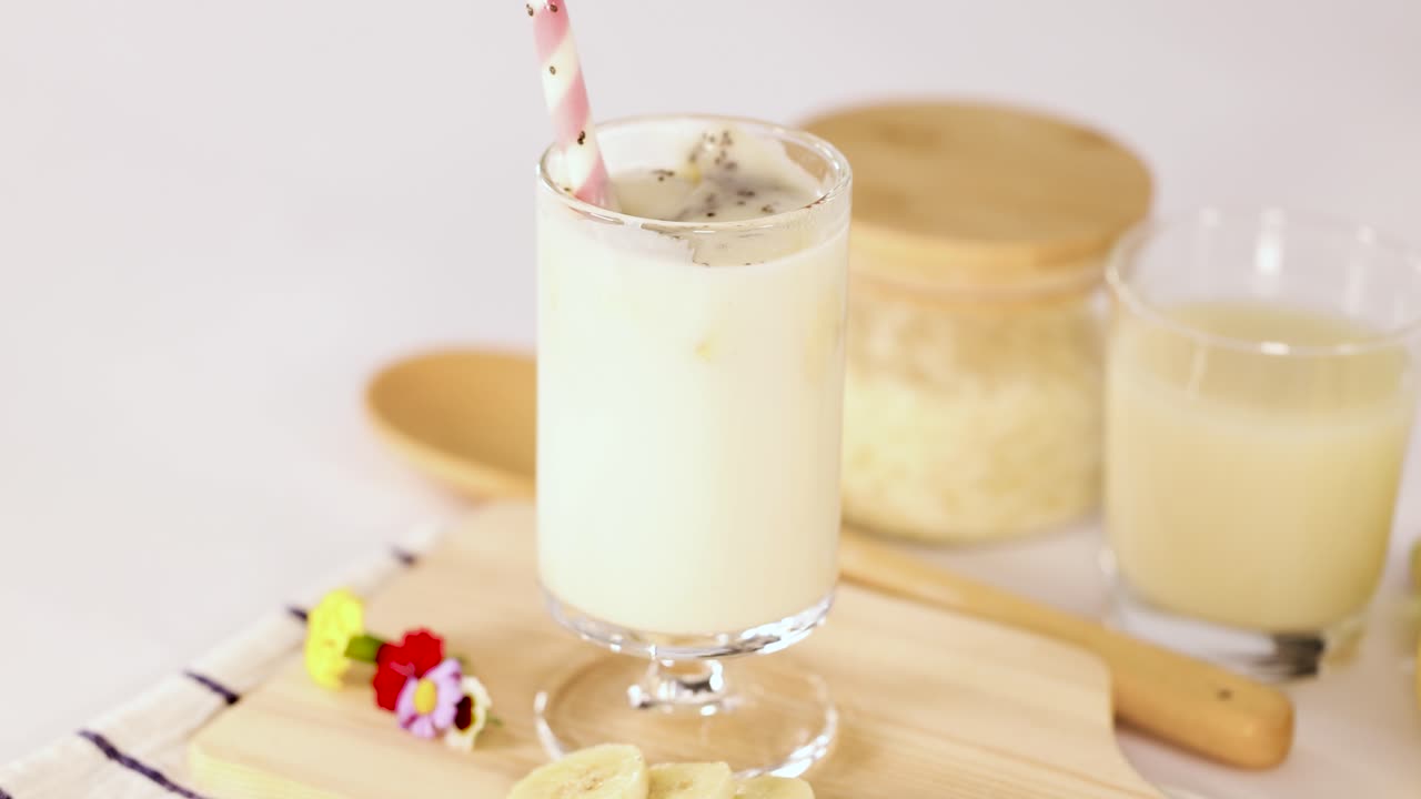 Hand stirs banana milkshake in glass, bright kitchen lighting, close-up, minimal camera movement