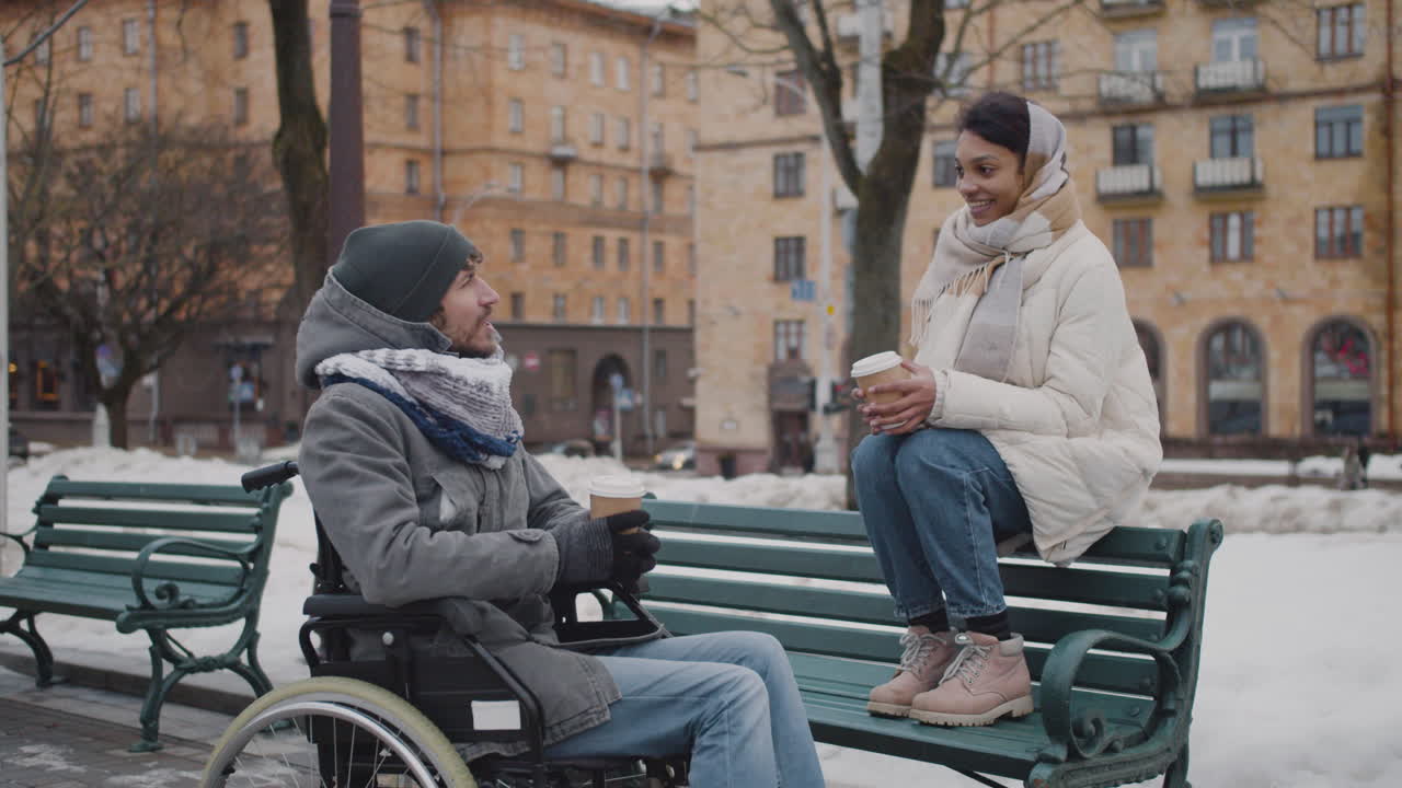 겨울에 도시의 벤치에서 테이크아웃 커피를 마시는 휠체어를 탄 이슬람 여성과 장애인 친구