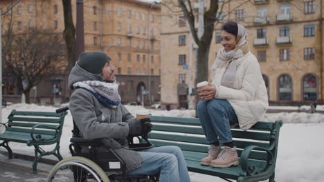 Mujer-Musulmana-Y-Su-Amiga-Discapacitada-En-Silla-De-Ruedas-Bebiendo-Café-Para-Llevar-En-Un-Banco-En-La-Ciudad-En-Invierno