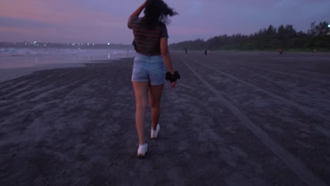 A-shot-following-behind-an-attractive-Asian-female-taking-a-twilight-walk-along-a-beach-at-low-tide-as-her-hair-flutters-in-the-evening-breeze,-Goa,-India