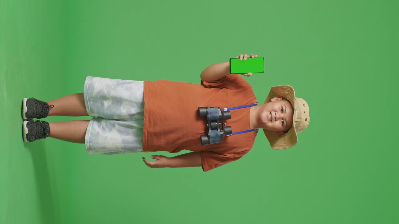 모자와 안경을 입은 아시아 관광 소년은 미소 짓고 카메라에 녹색 화면 스마트폰을 보여줍니다. 소년 연구원은 녹색 스크린 배경에서 무언가를 조사합니다.