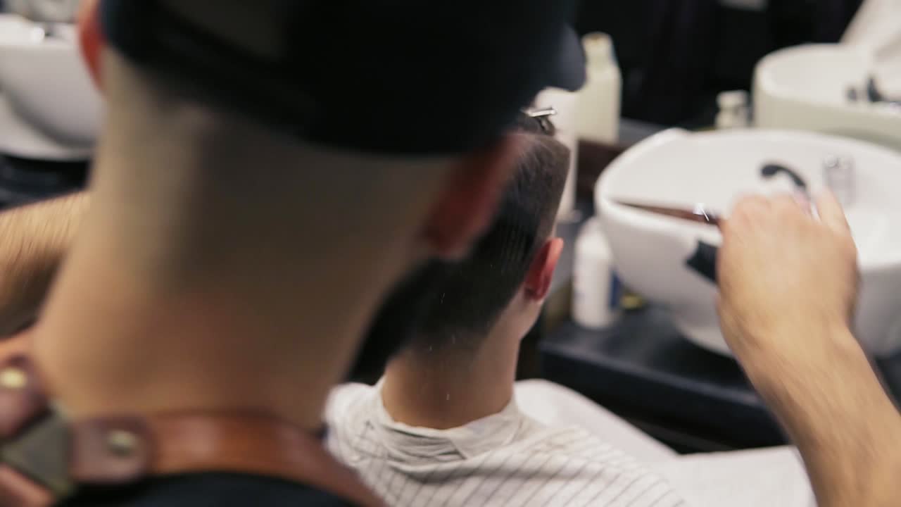 Back view of a client sitting by the mirror while a bearded barber is performing a haircut with scissors and combing his client