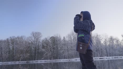 Niño-Con-Un-Abrigo-Azul,-Una-Bolsa-De-Hombro-Marrón-Parado-En-La-Orilla-Del-Río-En-Un-Paisaje-Nevado,-Mirando-A-Través-De-Binoculares