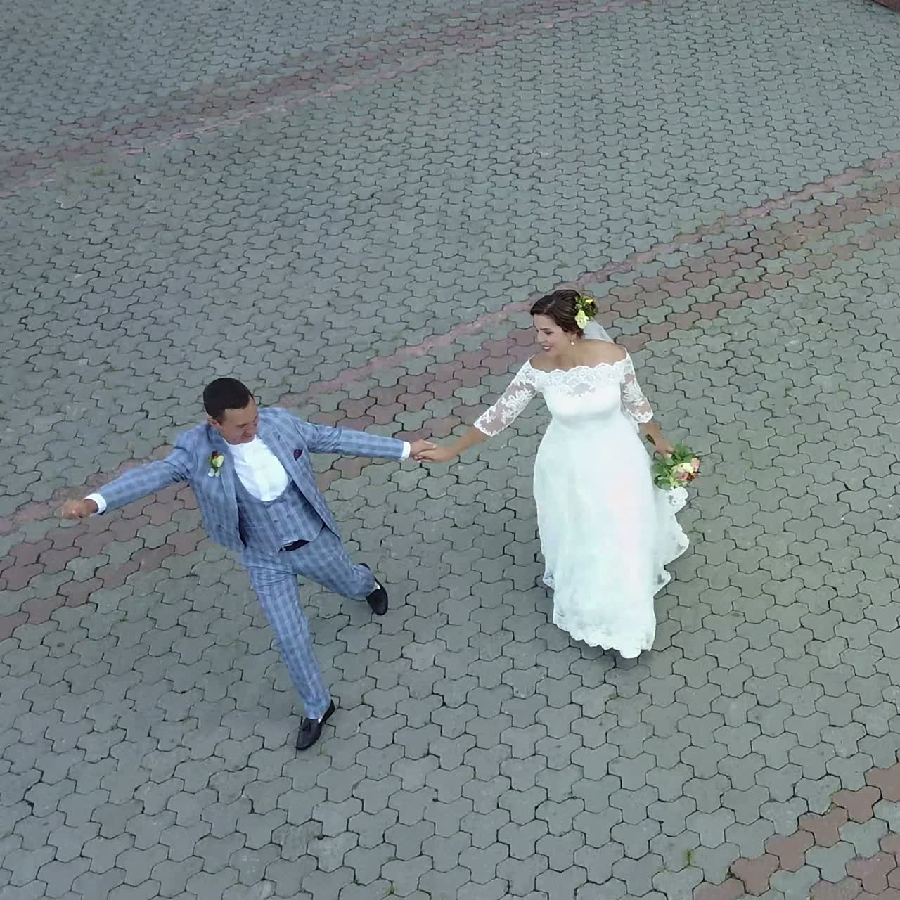 Charming stylish newlyweds are holding hands while walking along the sunny town street. Aerial view. Square video