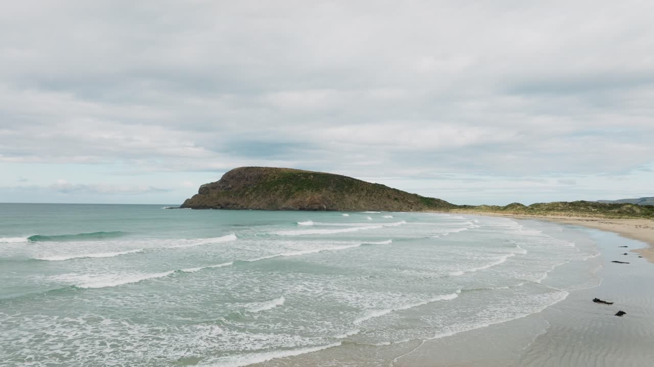 vista aérea sobre una remota playa de arena con olas ondulantes en aguas poco profundas del océano y la península de la bahía de caníbal en catlins, isla sur de nueva zelanda aotearoa