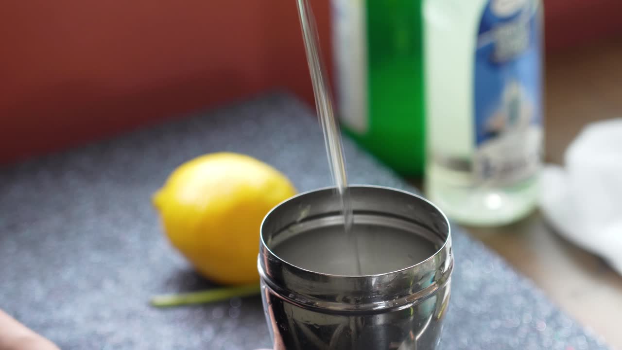 Bartender Pours Clear Liquid Vodka or Tequila into a Silver Cobbler Shaker for a Cocktail or Mixed Drink with Lemon, mint and Cucumber Slices, Closeup of Beverage with Pour Spout and Woman’s Hands