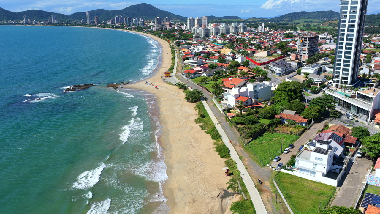 Top-down aerial tilt over breaking waves at Praia da Armação tilting up to reveal the Penha city skyline on a sunny day.