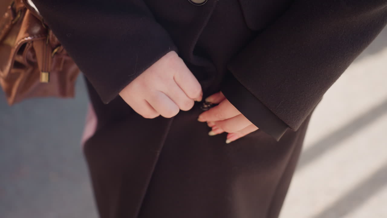 소녀가 손톱으로 똑딱 단추를 눌러 모직 코트를 여미고 있어요. 옆에는 갈색 가방이 있고, 햇빛이 옷감에 비치고 있네요. 손과 단추에 초점을 맞춘 겨울 스타일의 순간으로, 부드러운 바람과 깔끔하고 미니멀한 패션 디테일이 돋보여요.