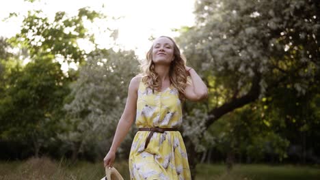 Fröhliche,-Selbstbewusste-Junge-Frau,-Die-Im-Sommerkleid-Mit-Strohhut-In-Der-Hand-Durch-Den-Grünen-Park-Geht