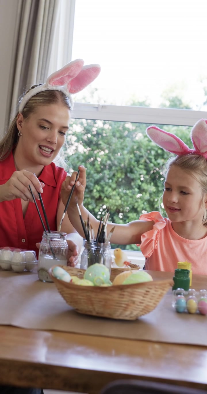 Vertical video: Mom joining left guiding girl painting Easter eggs at table by window, bunny ears