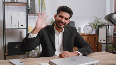 Hombre-De-Negocios-Trabajando-En-Una-Computadora-Portátil-Sonriendo-Amablemente-A-La-Cámara-Y-Saludando-Con-Las-Manos-En-La-Oficina