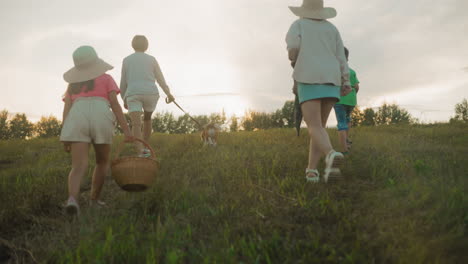 family walk on grassy hill with dog in warm sunset light, capturing movement and joy, young girl in hat leads with basket, others follow, creating a peaceful outdoor scene