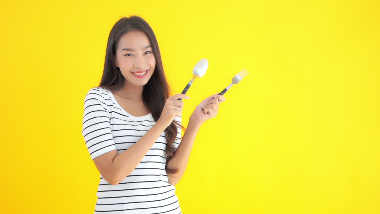 Portrait of Attractive Asian Female Pointing With Fork and Spoon on Right. Cooking and Kitchen Interactive Media Content, Isolated on Yellow Background